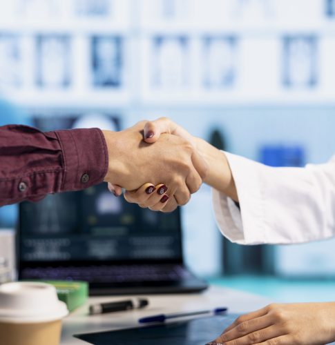Indian general practitioner greeting her male patient at a medical examination, sharing a handshake and discussing about diagnostic reports. Woman doctor helping man to treat an illness. Close up.